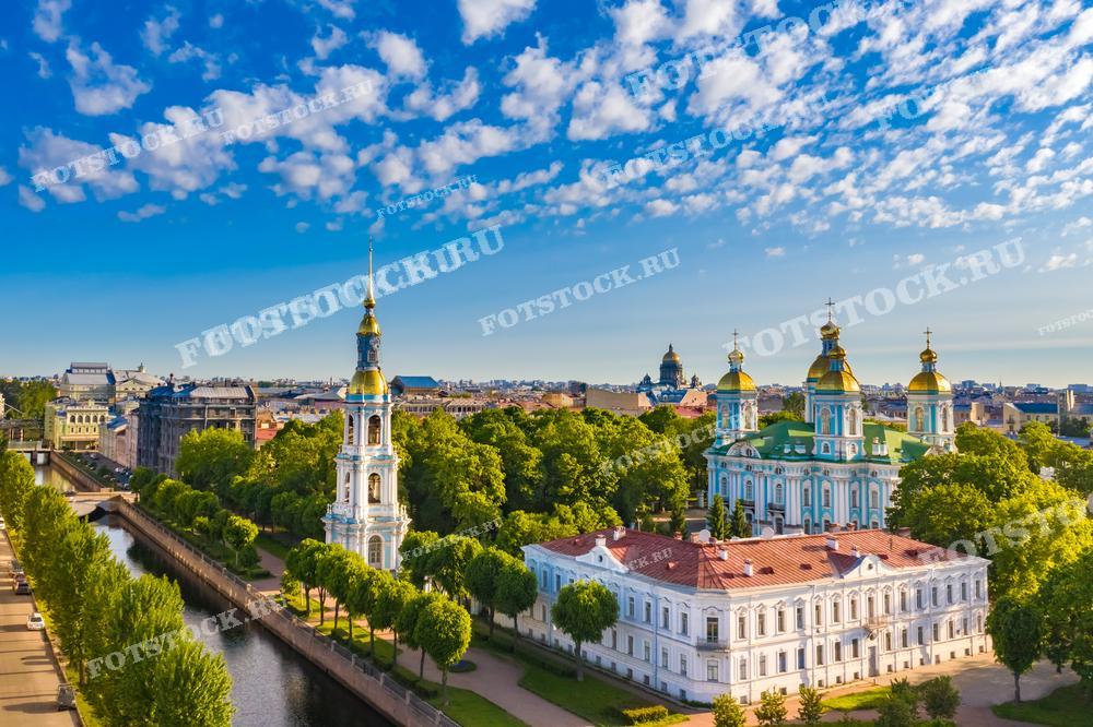 На фотографии изображён Никольский морской собор в Санкт-Петербурге на фоне ясного голубого неба с белыми облаками. Собор с золотистыми куполами возвышается над зелёной листвой деревьев, а рядом течёт канал с водной гладью, отражающей небесные тона. Это место является значимым культурным и историческим символом, фото подходит для использования в путеводителях, исторических обзорах, религиозных изданиях или как элемент декора, отражающий красоту русской православной архитектуры.